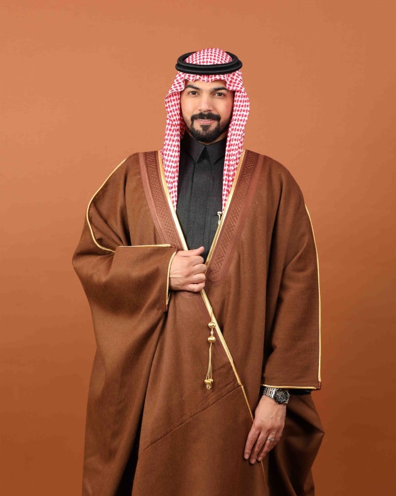 رجل يرتدي بشتاً شتوياً بلون أشقر مائل إلى البني بحواف ذهبية مع شماغ أحمر
